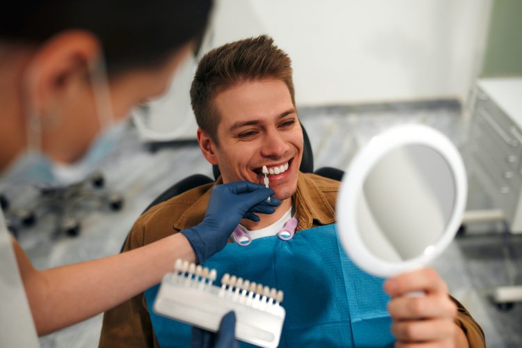 Medicine, dentistry and healthcare concept - closeup of a dentist with tooth color samples choosing a shade for a male patient's teeth in a dental clinic looking at a mirror.